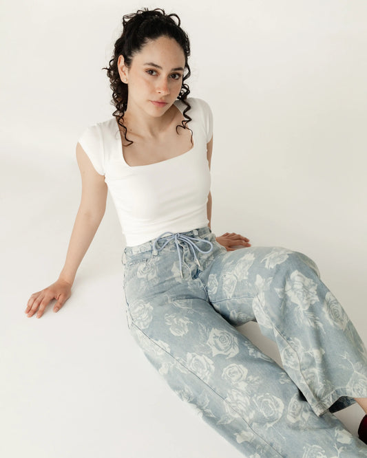 Mujer joven con blusa blanca y jeans claros estampados de rosas, sentada en fondo blanco