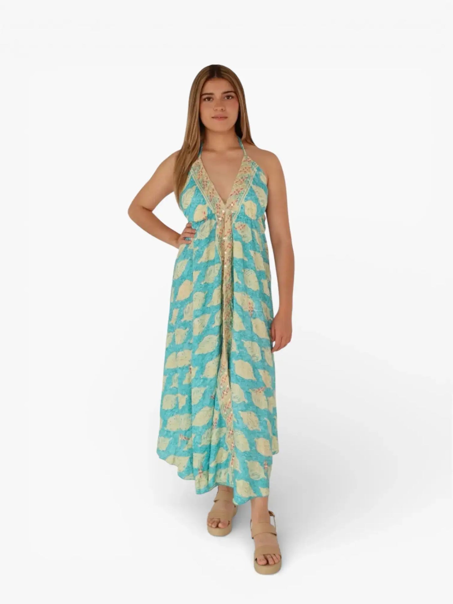 Mujer joven con vestido largo azul estampado, escote en V y sandalias beige, fondo blanco.