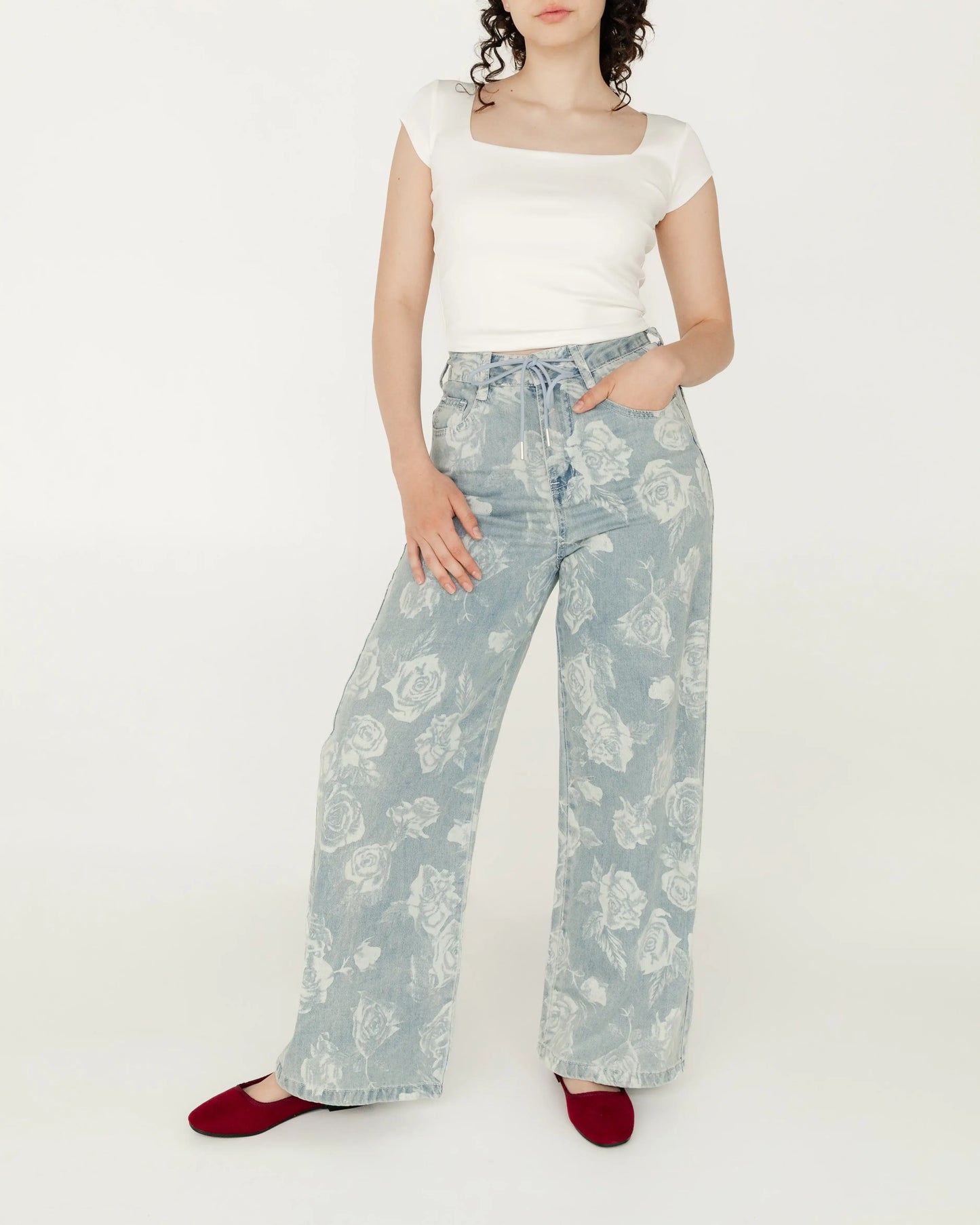 Mujer con playera blanca corta y pantalón de mezclilla azul claro con estampado floral, zapatos rojos.