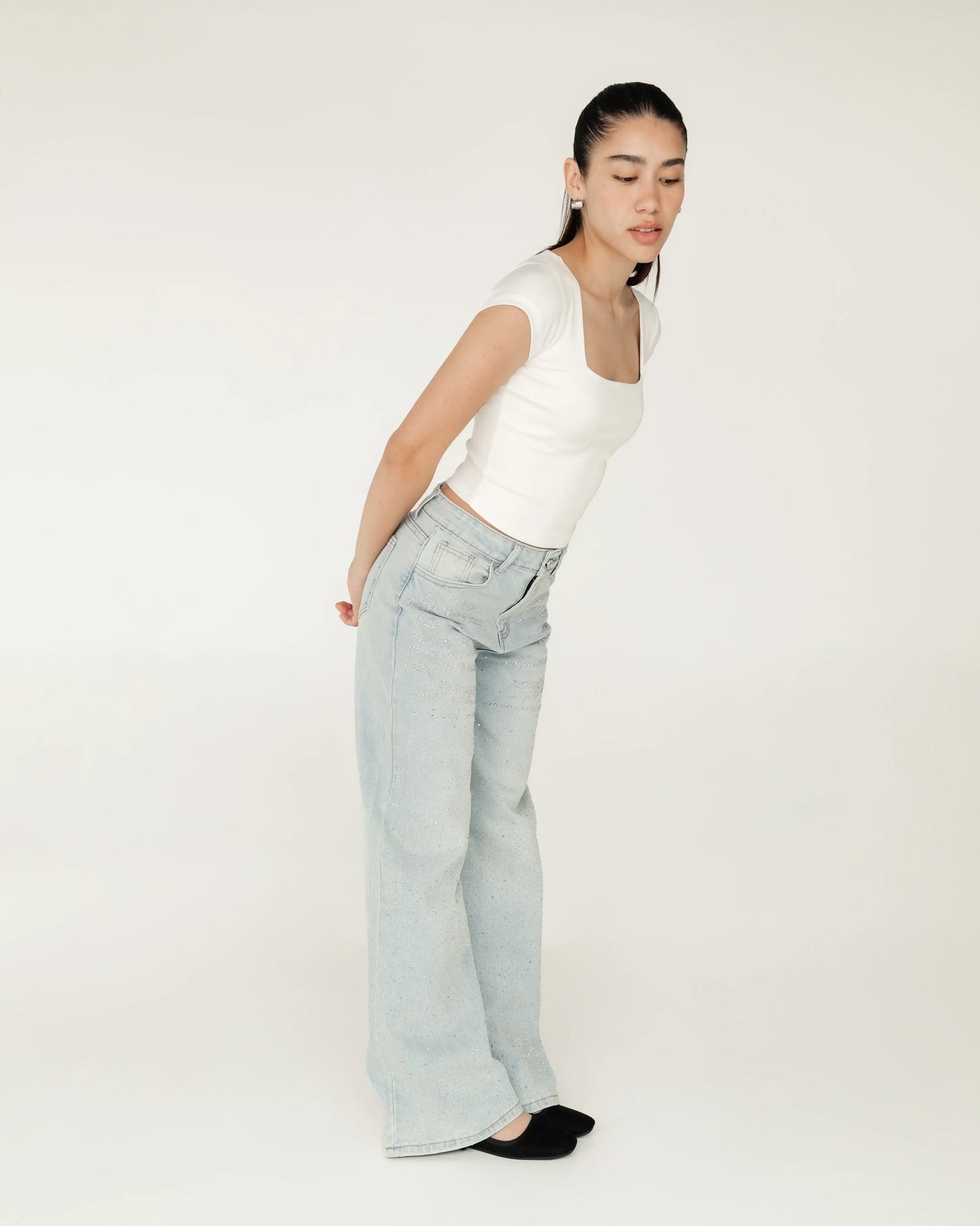 Mujer joven con top blanco y jeans claros holgados, posando en fondo blanco minimalista.