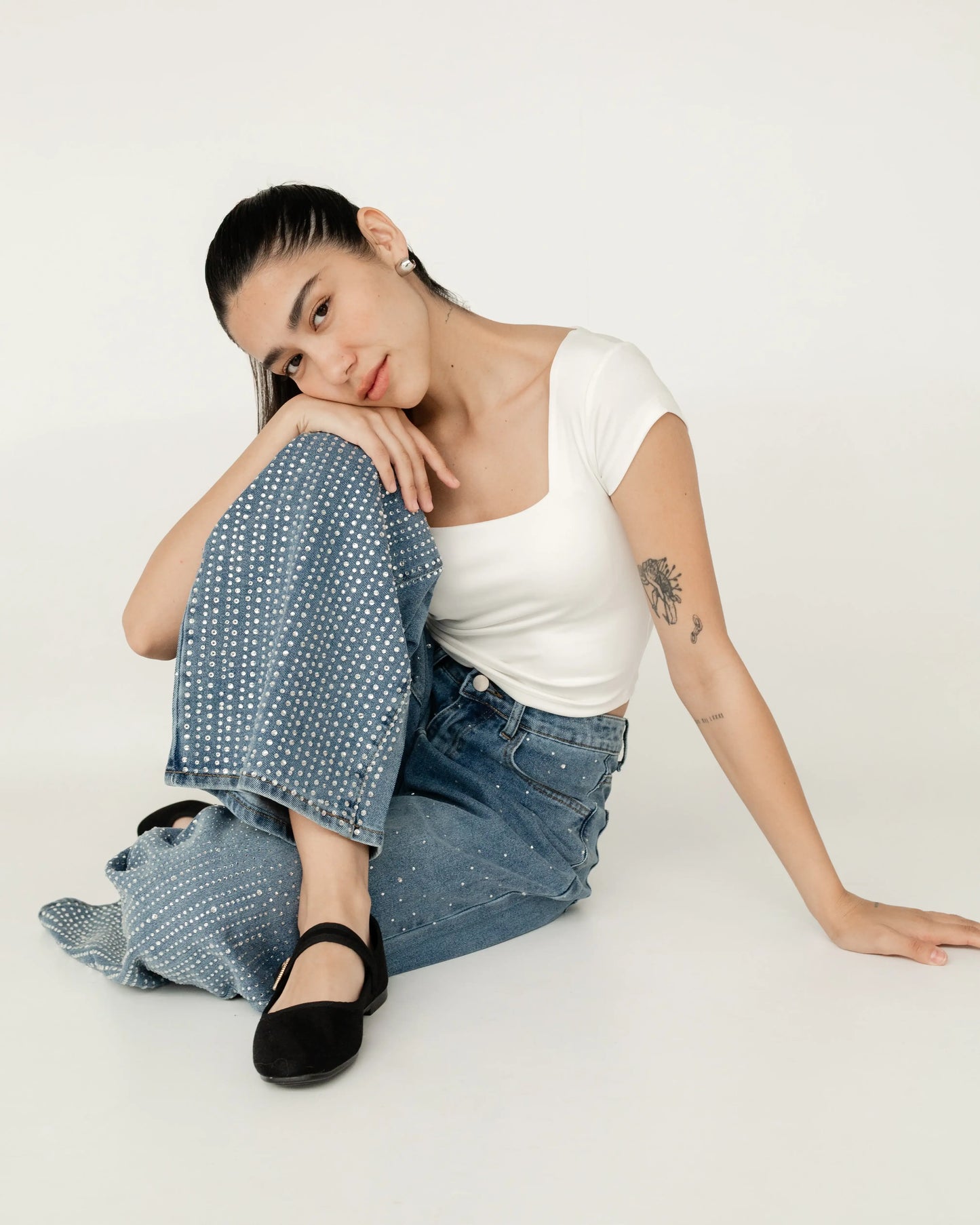 Mujer sentada con blusa blanca, jeans de mezclilla azul con perlas y zapatos negros