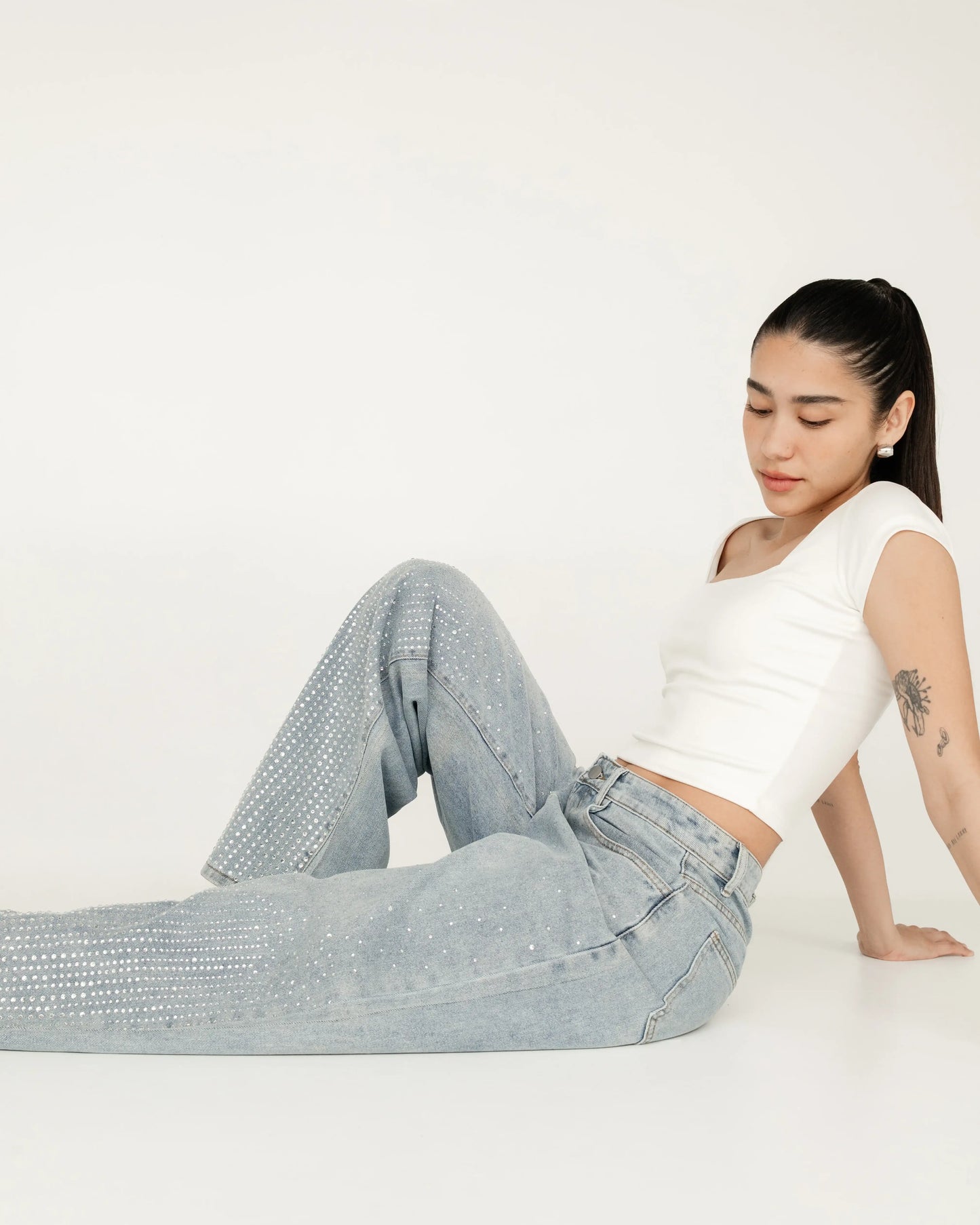 Mujer joven con top blanco y jeans claros con brillos, posando en fondo blanco minimalista.
