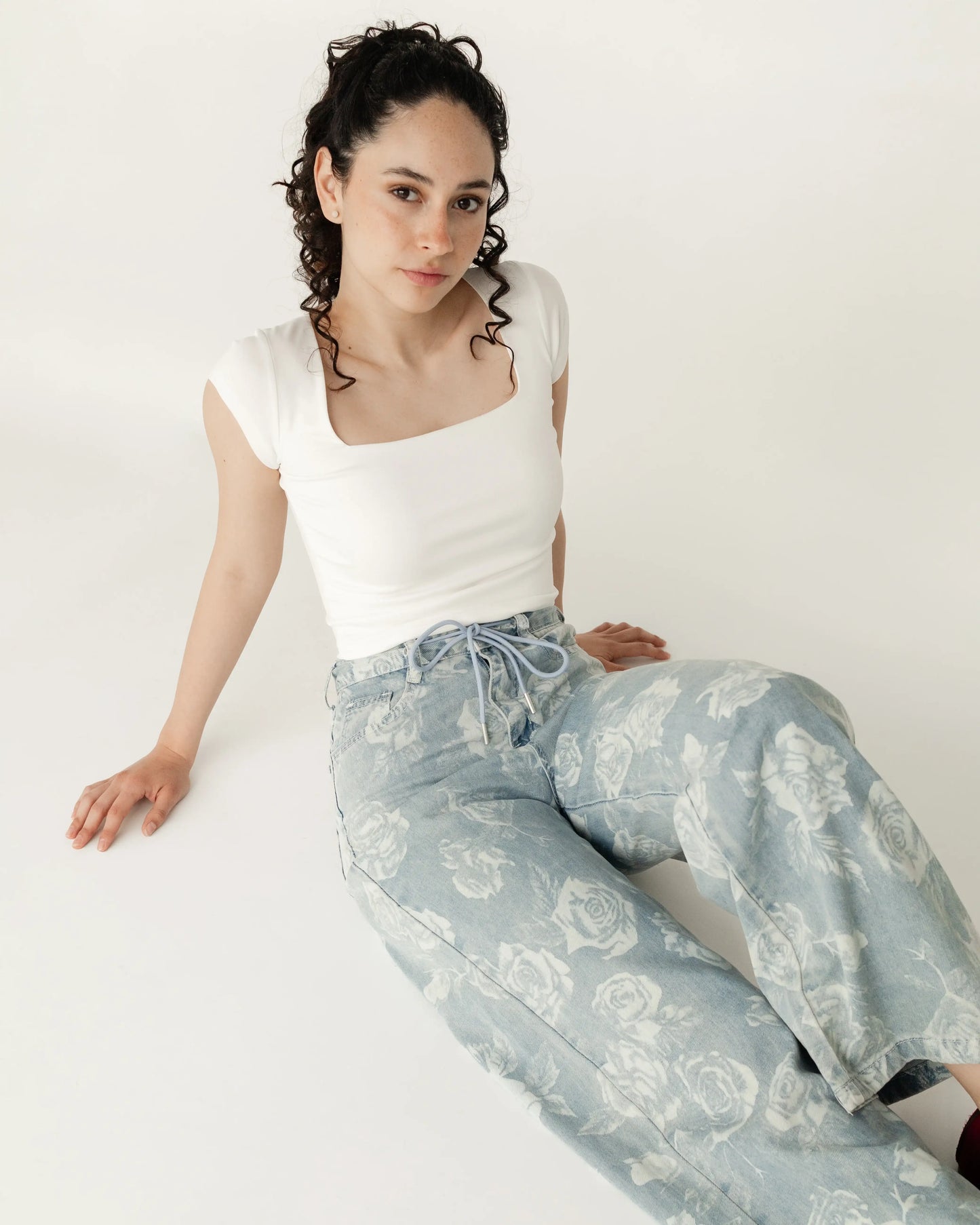 Mujer joven con blusa blanca y jeans claros estampados de rosas, sentada en fondo blanco