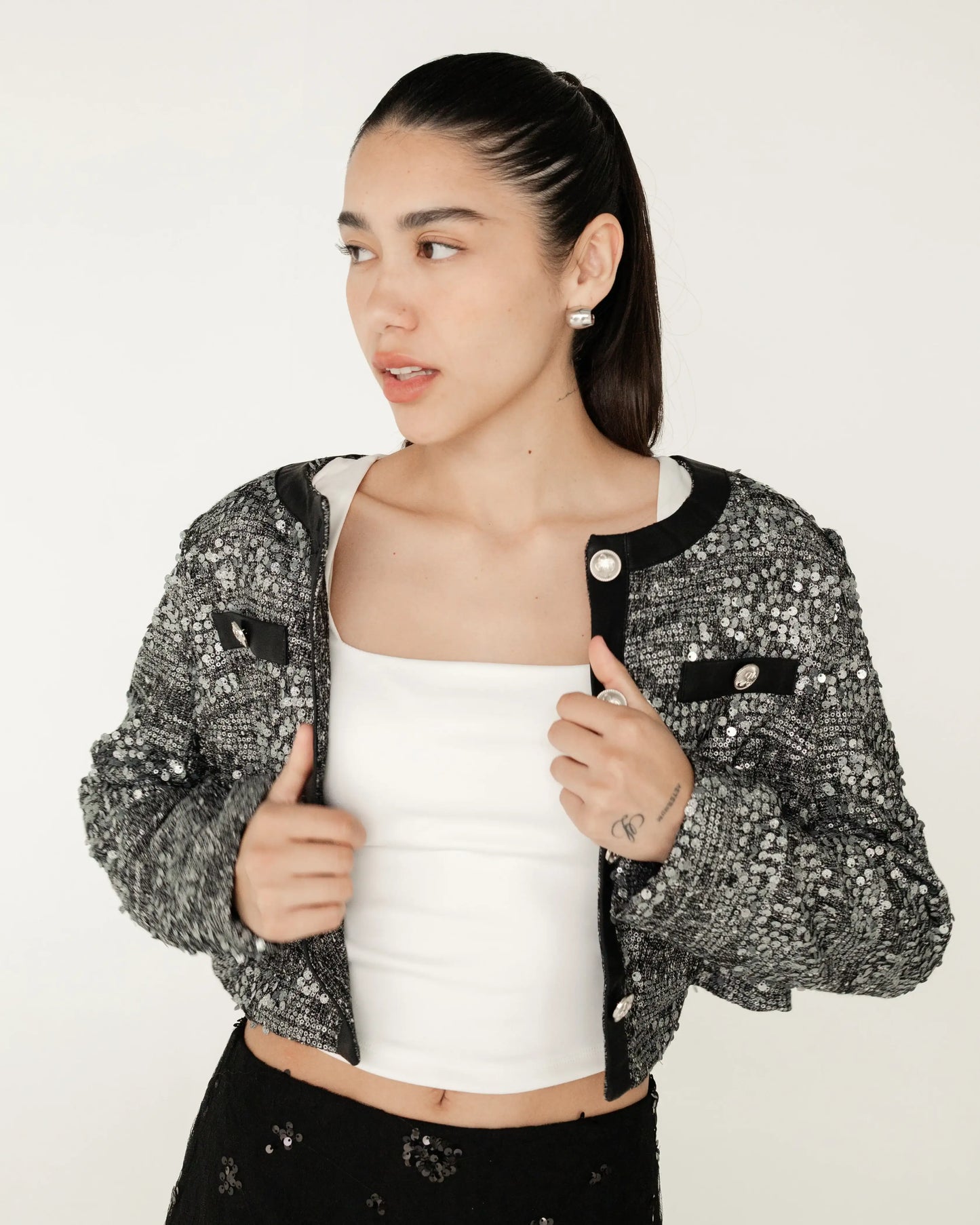 Mujer joven con saco tweed negro y plata, top blanco y pantalón negro, fondo neutro