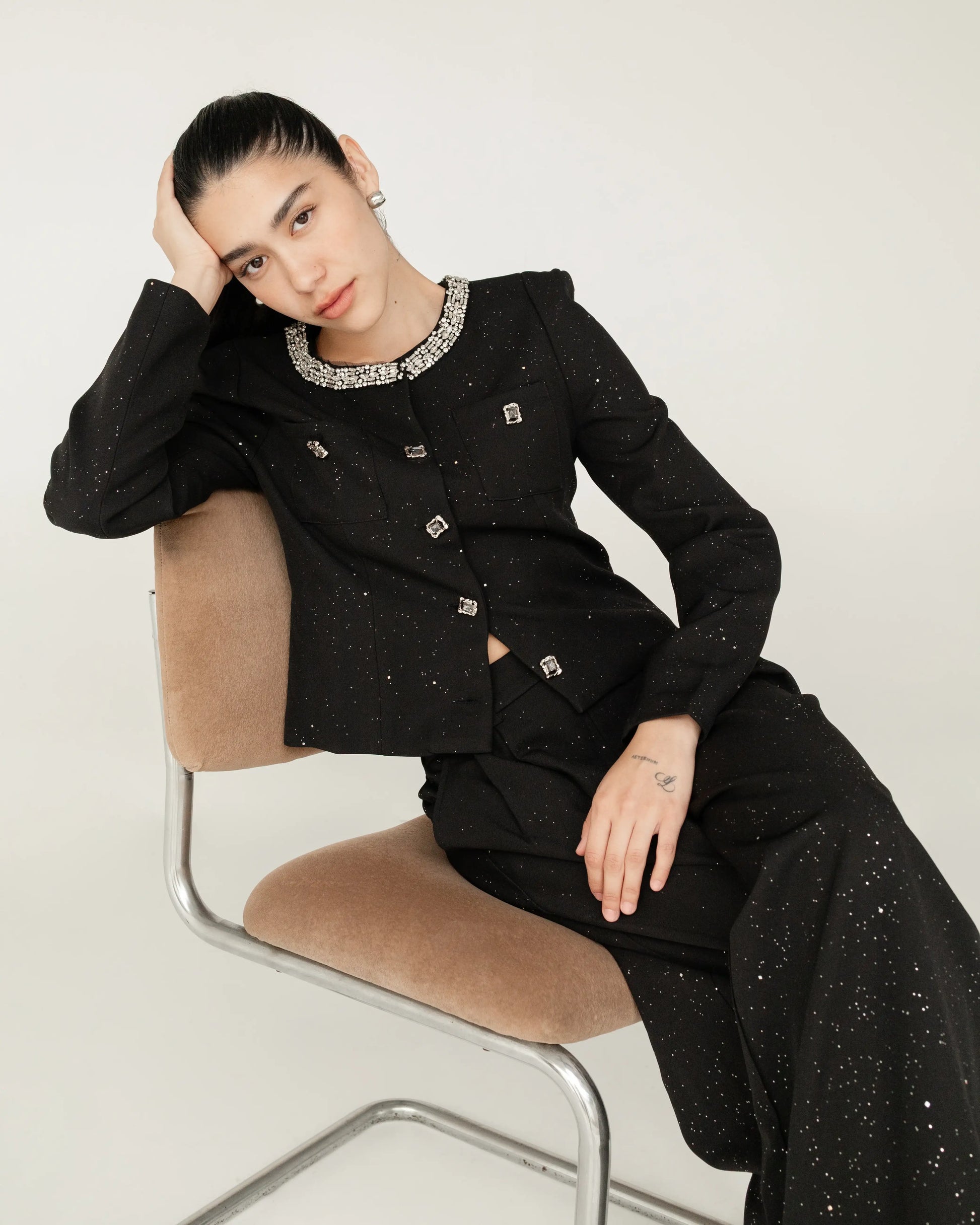 Mujer con saco y pantalón negro con detalles brillantes, sentada en una silla, estilo elegante.