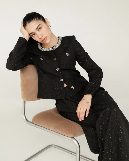 Mujer con saco y pantalón negro con detalles brillantes, sentada en una silla, estilo elegante.