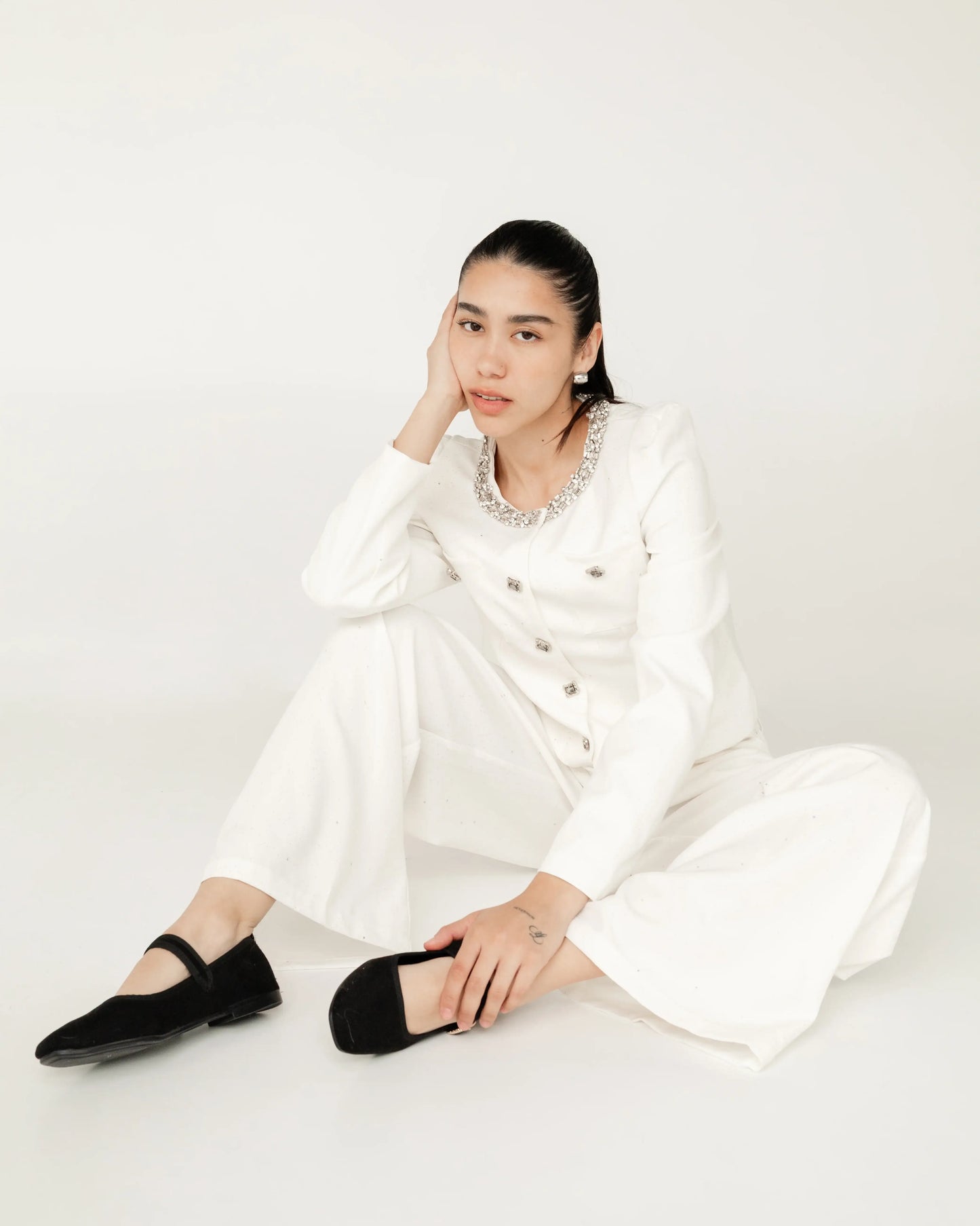 Mujer joven sentada con conjunto blanco elegante y zapatos negros, fondo minimalista